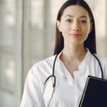 Crop smiling young ethnic female doctor in medical uniform with stethoscope and notebook standing in modern medical room with panoramic window