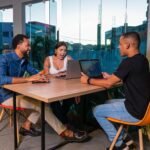 Three colleagues sitting at a table working on laptops in a vibrant, urban office space.